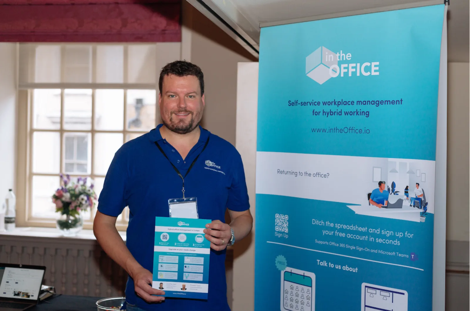 Jon Kent in an intheOffice polo shirt holding a leaflet beside the intheOffice roll-up banner at the Office Management Show