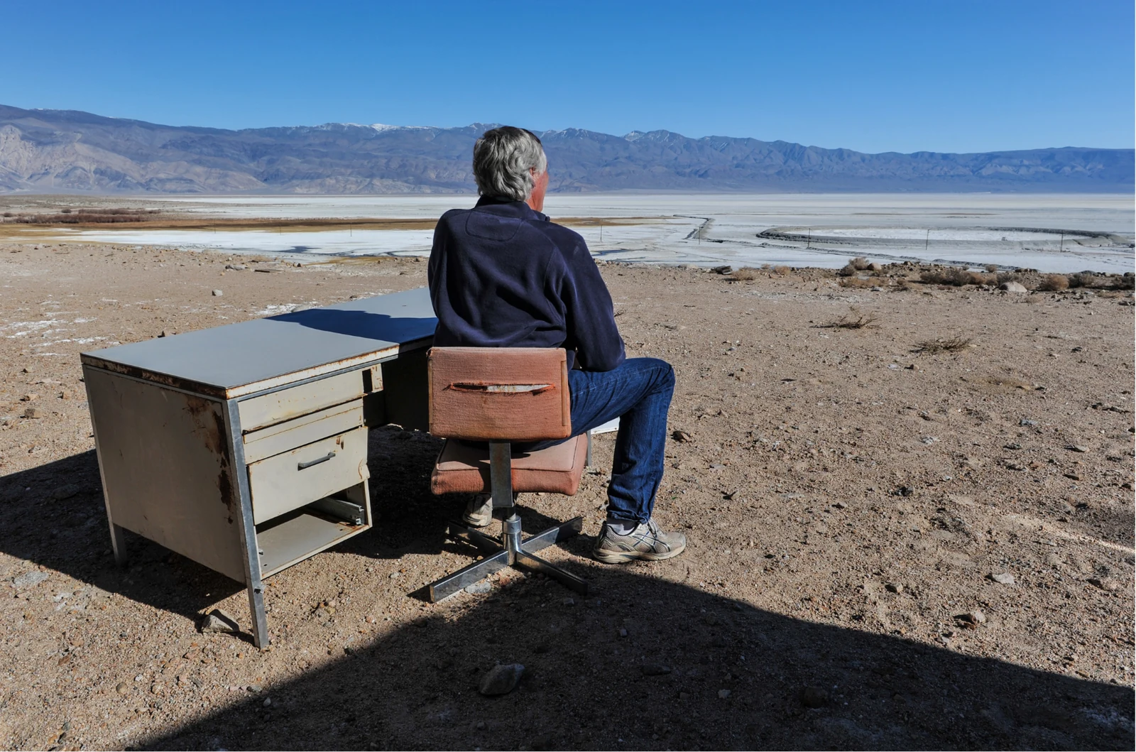 A man sitting alone at an old office desk in the middle of a desert